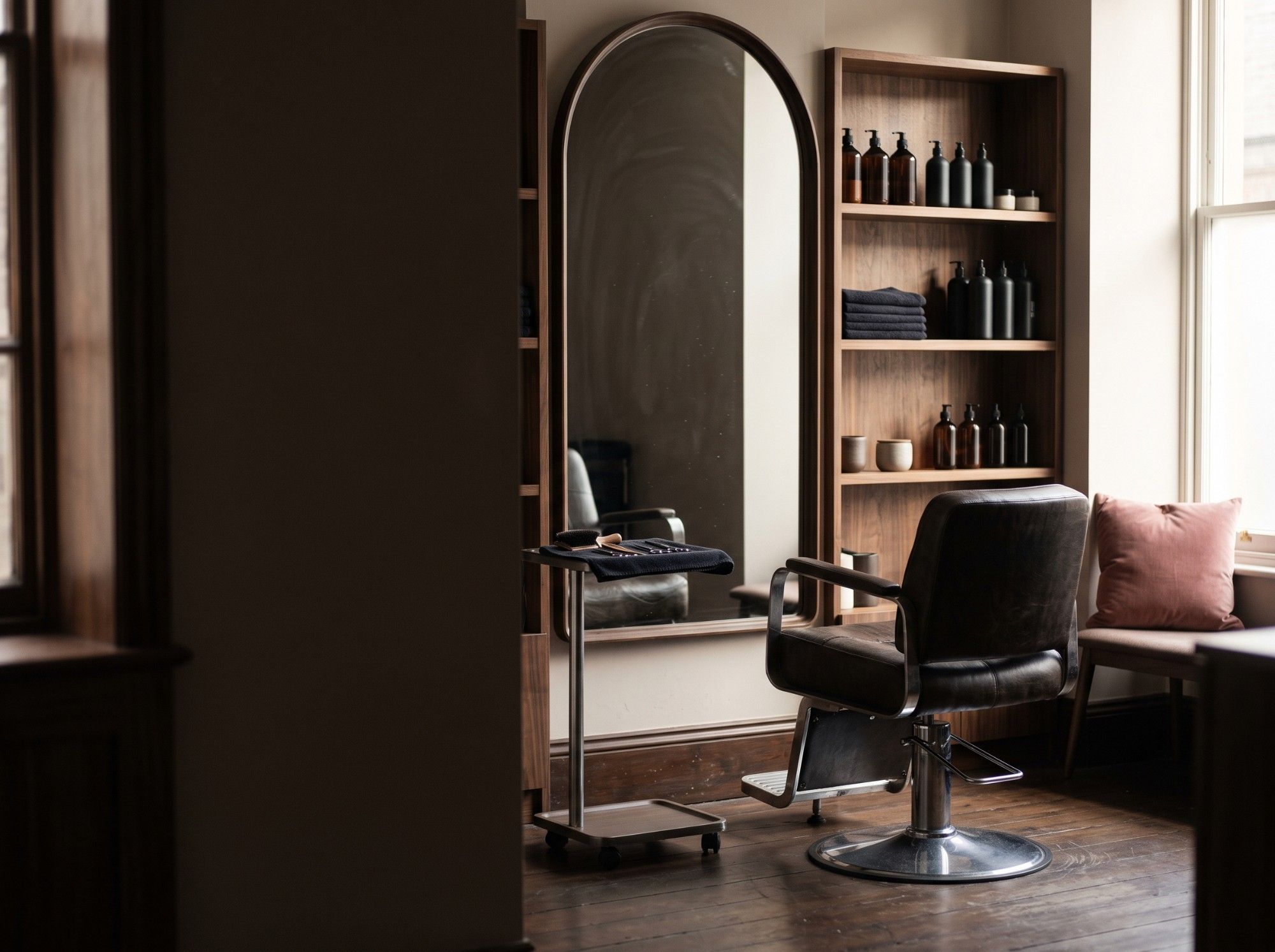 A calm hair studio with an arched mirror, a leather styling chair, walnut shelves and a rose pink cushion by the window.