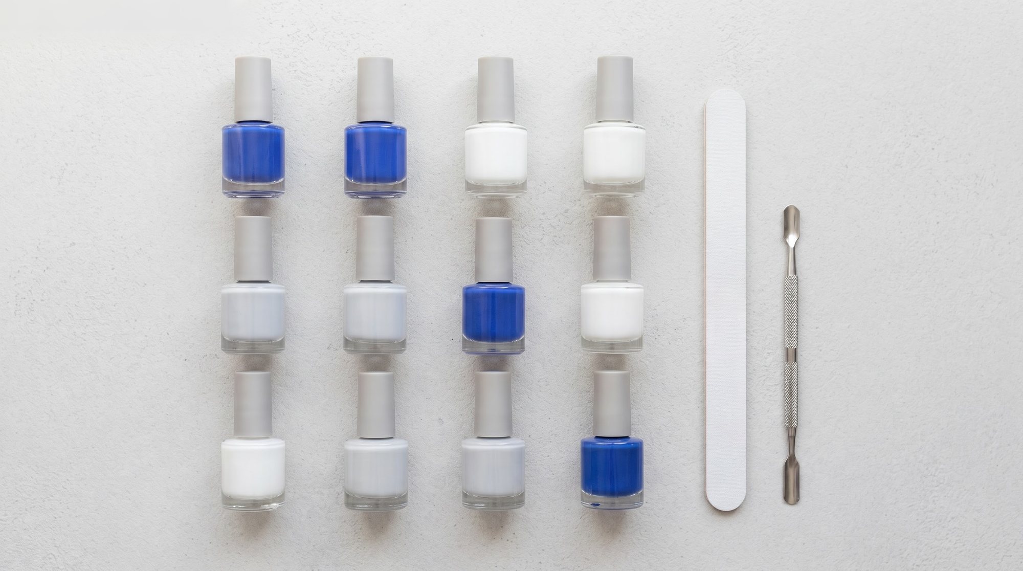 Rows of nail polish bottles in cobalt blue, white and pale grey, with a nail file and a cuticle tool, on a light grey surface.